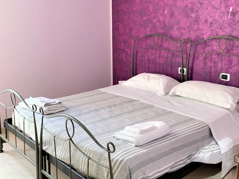 Una camera dalle pareti viola e dal letto con struttura in metallo, perfetta per un soggiorno rilassante presso le case vacanze a Lampedusa.