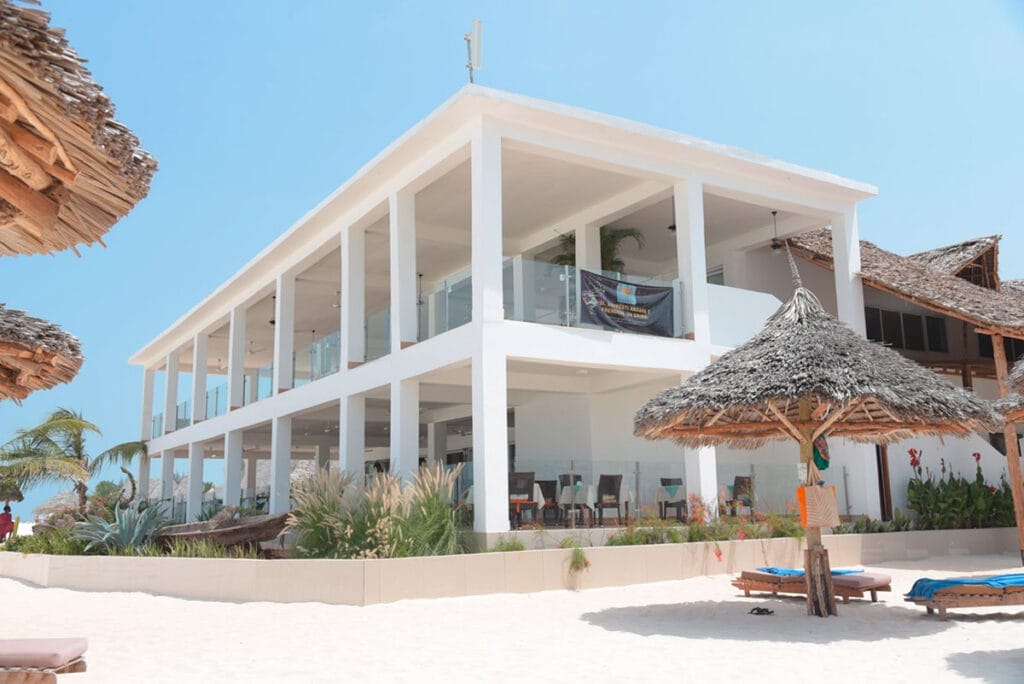 Un edificio bianco con ombrelloni in paglia sulla spiaggia, che offre un pacchetto viaggio a Zanzibar di SBS Viaggi.