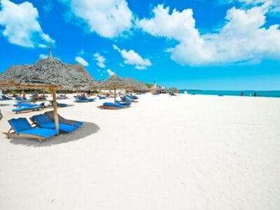Una bellissima spiaggia di sabbia bianca ornata da sedie a sdraio e ombrelloni blu. Questo paradiso da cartolina fa parte di un pacchetto di viaggio esclusivo offerto da Bravo Premium Kendwa Beach, disponibile tramite SBS Viaggi.