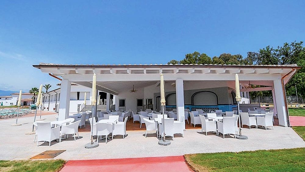 Patio esterno con zona pranzo con tavoli e sedie bianchi sotto un pergolato, adiacente ad una piscina con vista in lontananza su Marina Torre Navarrese.