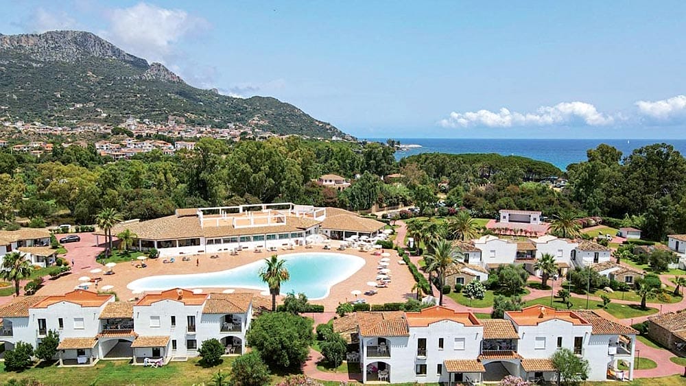 Una località costiera con porto turistico e piscina circondata da edifici bianchi e vegetazione lussureggiante, con le montagne sullo sfondo.