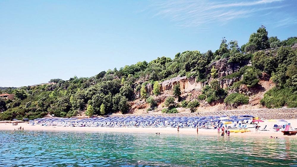 Spiaggia con lettini blu, visitatori e verde circostante in una giornata di sole al Club Esse Cala Gonone, Sardegna.