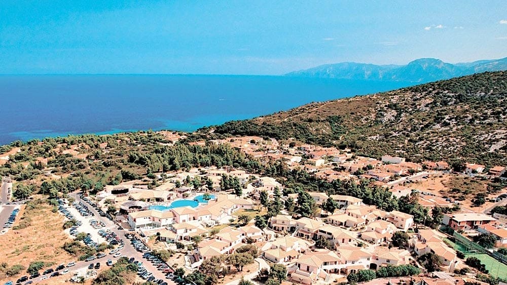 Veduta aerea del Club Esse Cala Gonone, un villaggio costiero della Sardegna con edifici dai tetti rossi e una piscina vicino a un mare cristallino.