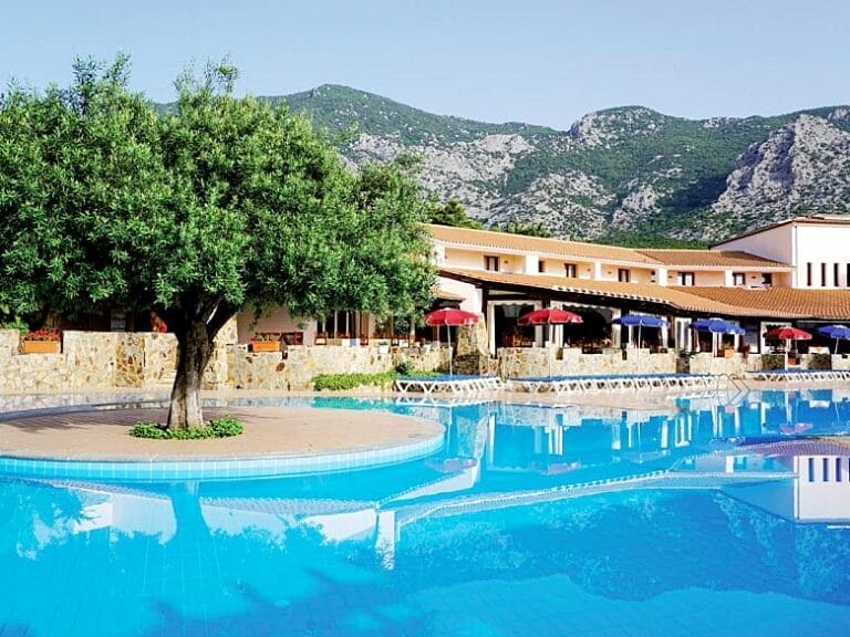 La tranquilla area piscina dell'hotel Club Esse con un albero, ombrelloni e uno sfondo montuoso a Cala Gonone, in Sardegna.
