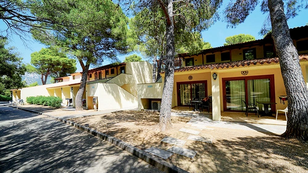 Villa in stile mediterraneo immersa tra i pini del Club Esse Palmasera Resort con facciata soleggiata e area salotto all'aperto in Sardegna.
