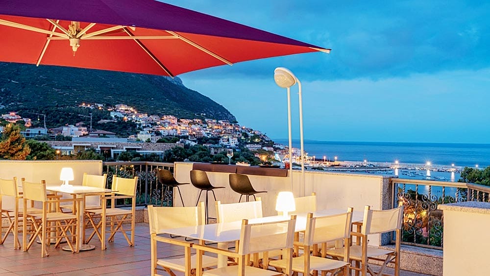 Zona pranzo all'aperto che si affaccia su una città costiera al tramonto con un grande ombrellone rosso e le luci della città in lontananza, immersa nel Resort Club Esse Palmasera, Sardegna.