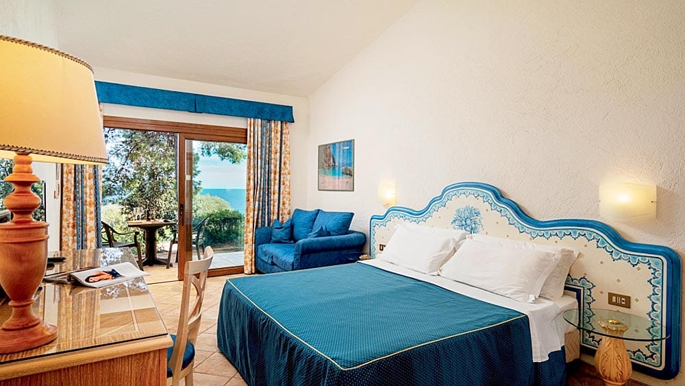Luminosa camera d'albergo al Club Esse Palmasera Resort con due letti, accenti di arredamento blu e un balcone con posti a sedere all'aperto.
