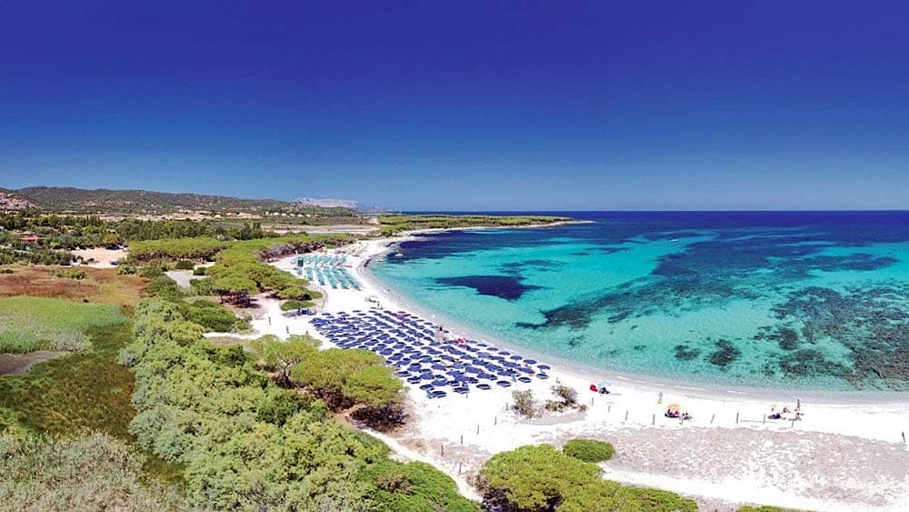 Veduta aerea di Eurovillage Sardegna con acque turchesi, spiaggia sabbiosa con ombrelloni e costruzioni nell'entroterra immerse nel verde.
