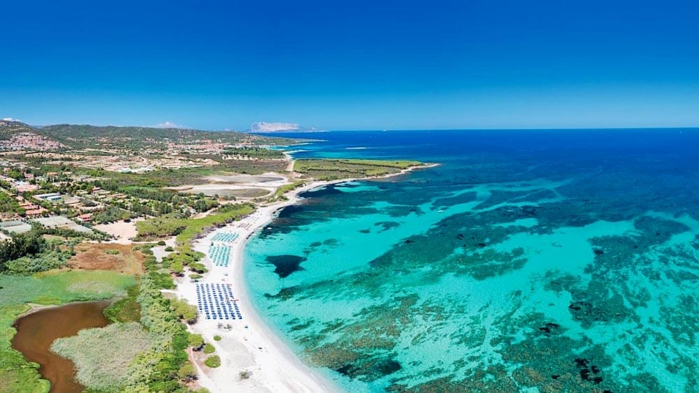 Veduta aerea di Eurovillage Sardegna con acque turchesi, spiaggia sabbiosa con ombrelloni e costruzioni nell'entroterra immerse nel verde.
