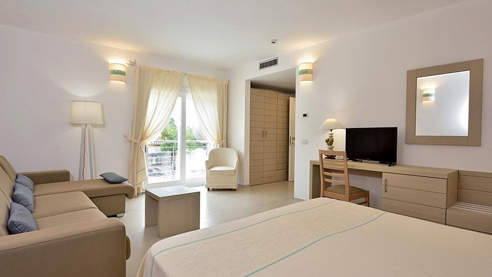 Camera d'albergo luminosa e moderna all'Eurovillage Sardegna con un ampio letto, un'area salotto, una TV e porte scorrevoli su un balcone.