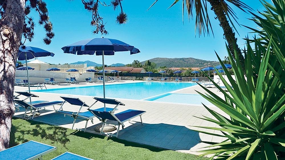 Una piscina all'aperto del resort Eurovillage in Sardegna con lettini e ombrelloni, incorniciata da alberi sotto un cielo azzurro e limpido.