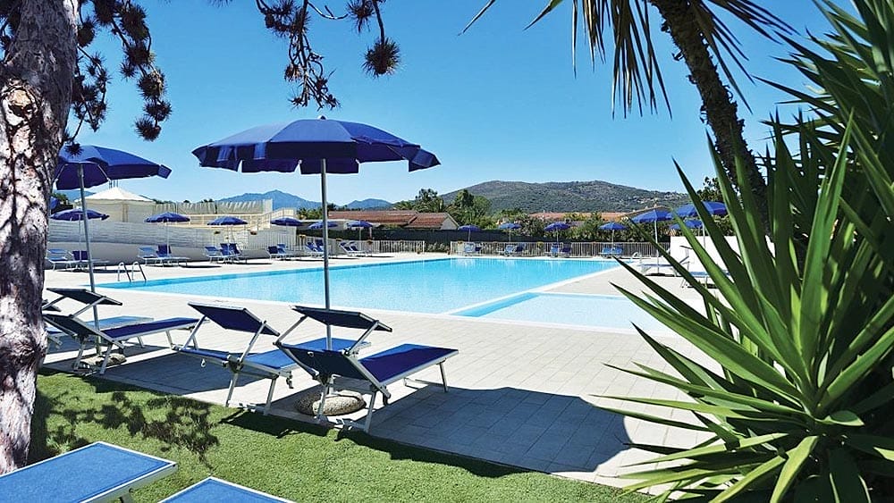 Un'assolata zona bordo piscina dell'Eurovillage Sardegna, con lettini e ombrelloni blu, immersa nel verde con vista sulle colline in lontananza.