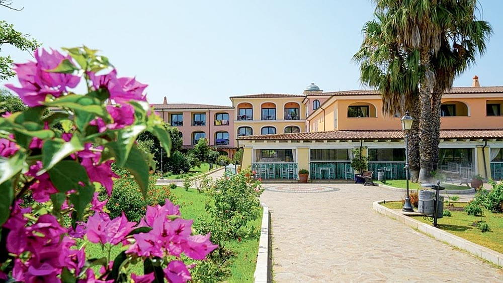 Un sentiero paesaggistico che conduce ad un hotel color terracotta con bouganville in primo piano al Garden Beach Sardegna.