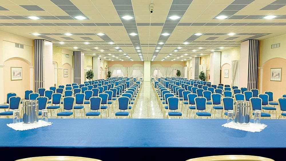 Una sala conferenze vuota al Marina Resort con file di sedie blu e un palco nella parte anteriore.