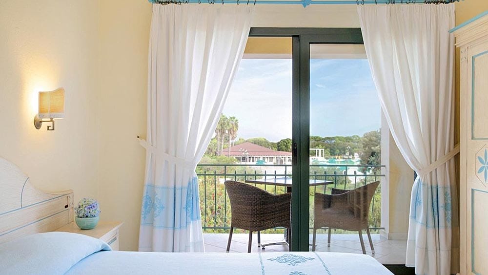 Camera da letto luminosa e ariosa con una porta finestra aperta che conduce a un'area salotto all'aperto del Garden Beach Sardegna, che offre una vista panoramica.