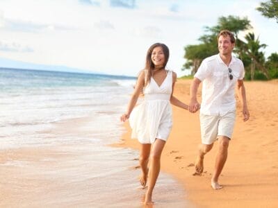 Una coppia felice che si tiene per mano e cammina lungo una spiaggia sabbiosa, con le onde dell'oceano che si infrangono dolcemente sulla riva accanto a loro, durante il loro viaggio di nozze.