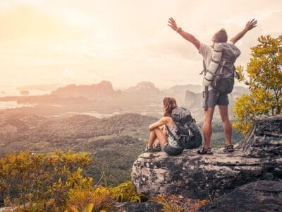 Due viaggiatori su una cima di montagna, uno seduto e contemplando l'orizzonte, l'altro in piedi con le braccia alzate durante il loro viaggio su misura di SBS Viaggi.