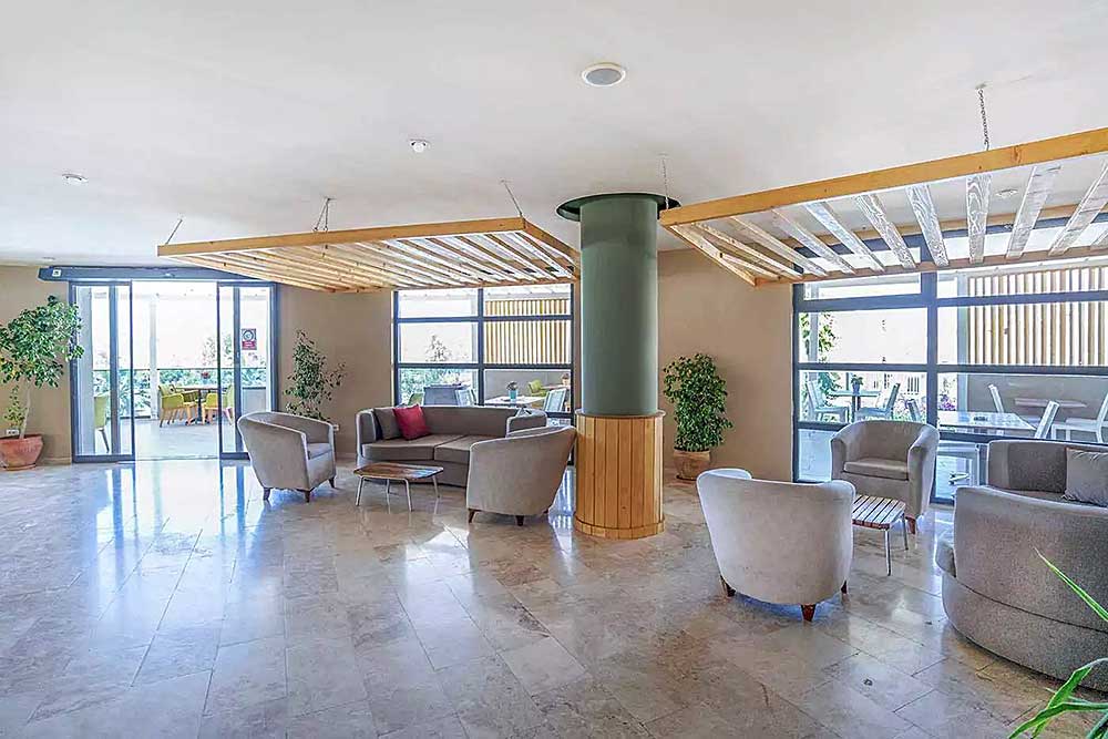 Lobby luminosa e moderna con poltrone grigie, porte in vetro, piante e soffitto in legno vicino alla spiaggia di Mare Bodrum.