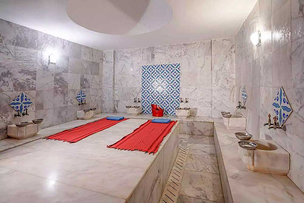 Un tradizionale hammam turco con interni in marmo, caratterizzato da una piattaforma centrale rialzata, tappetini rossi, piastrelle blu decorative e vasche di metallo vicino alla spiaggia di Mare Bodrum.