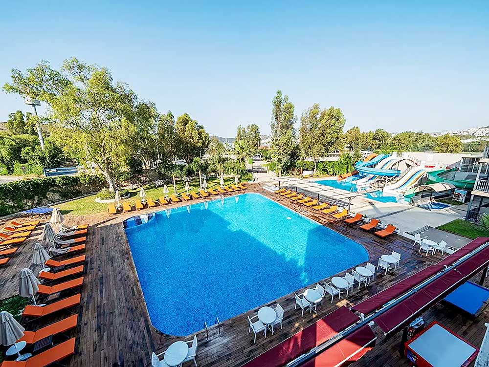 Area piscina all'aperto con sedie a sdraio e scivoli, circondata da alberi sotto un cielo azzurro sulla spiaggia di Mare Bodrum.