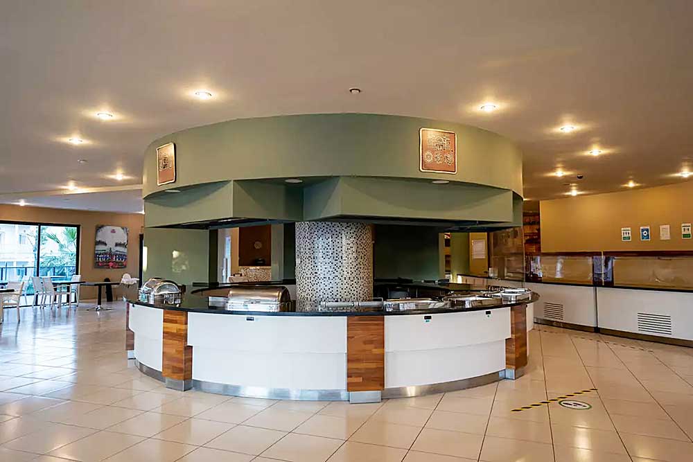 Una spaziosa caffetteria con una grande postazione circolare per il servizio di cibo al centro, dotata di attrezzature da cucina e banconi puliti, situata in uno spazio calorosamente illuminato sulla spiaggia di Bodrum.