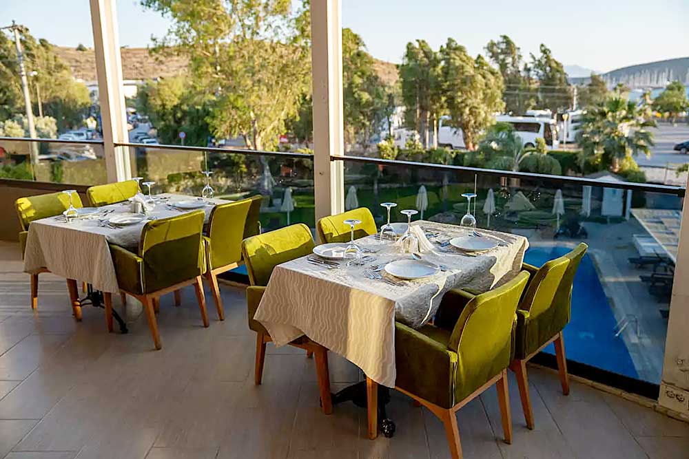 Elegante allestimento per pranzi all'aperto con tavoli rivestiti in lino bianco, apparecchiati con piatti e bicchieri, con vista sulla spiaggia di Mare Bodrum.