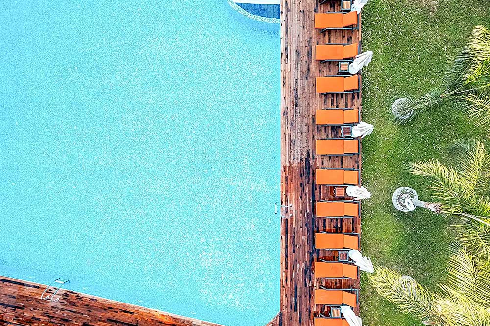 Veduta aerea di una grande piscina circondata da lettini e palme, con una terrazza in legno su un lato sulla spiaggia di Mare Bodrum.