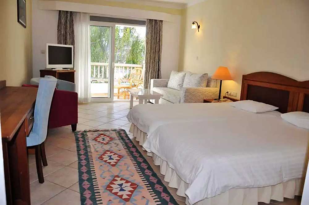 Luminosa camera dell'Hotel Okaliptus dotata di letto matrimoniale, piccola area salotto vicino alla finestra, TV su supporto e tappeto colorato su pavimento piastrellato.