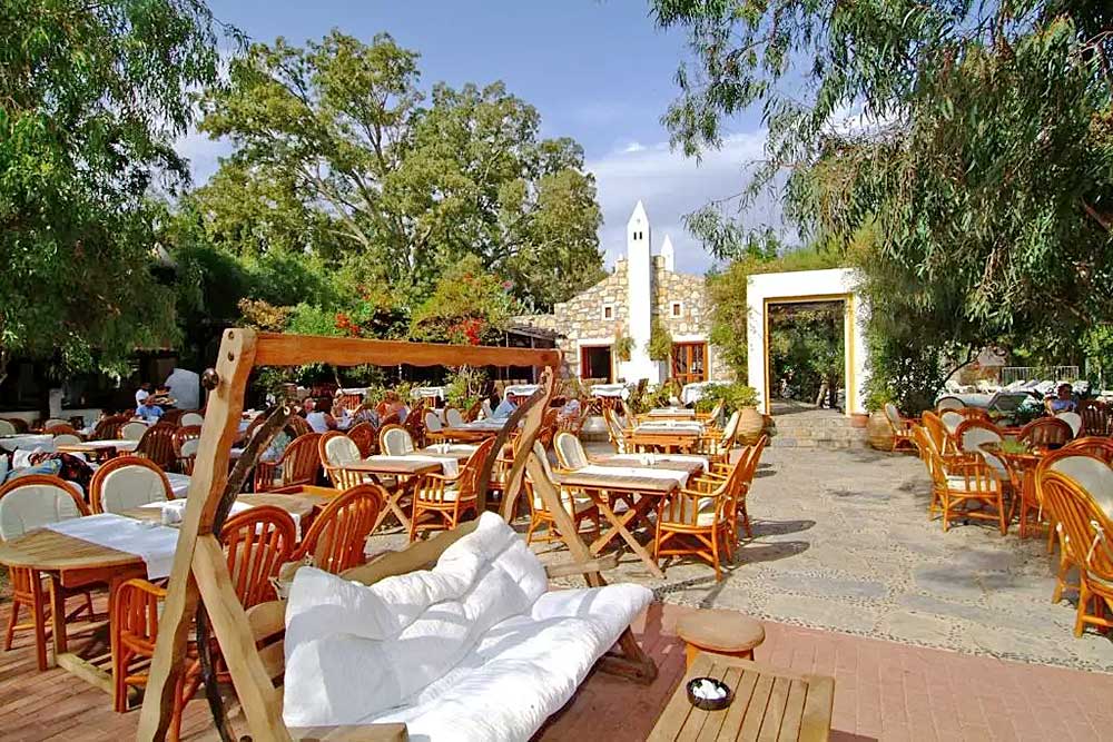 Patio esterno del ristorante con sedie e tavoli in vimini, divani bianchi sotto gli alberi e vista sullo storico edificio dell'Hotel Okaliptus con campanile.