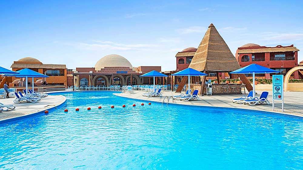 La piscina del resort Villaggio Mar Rosso con sedie a sdraio, ombrelloni e architettura a tema egiziano sotto un cielo azzurro e limpido.