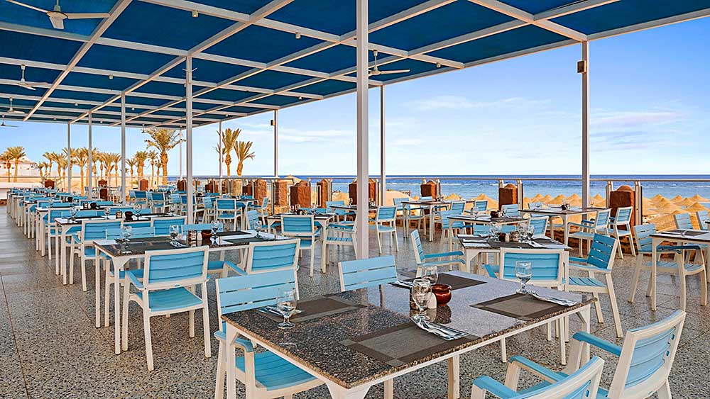 Ristorante sulla spiaggia con sedie e tavoli blu vuoti sotto una tettoia al Villaggio Mar Rosso, con vista sul mare.