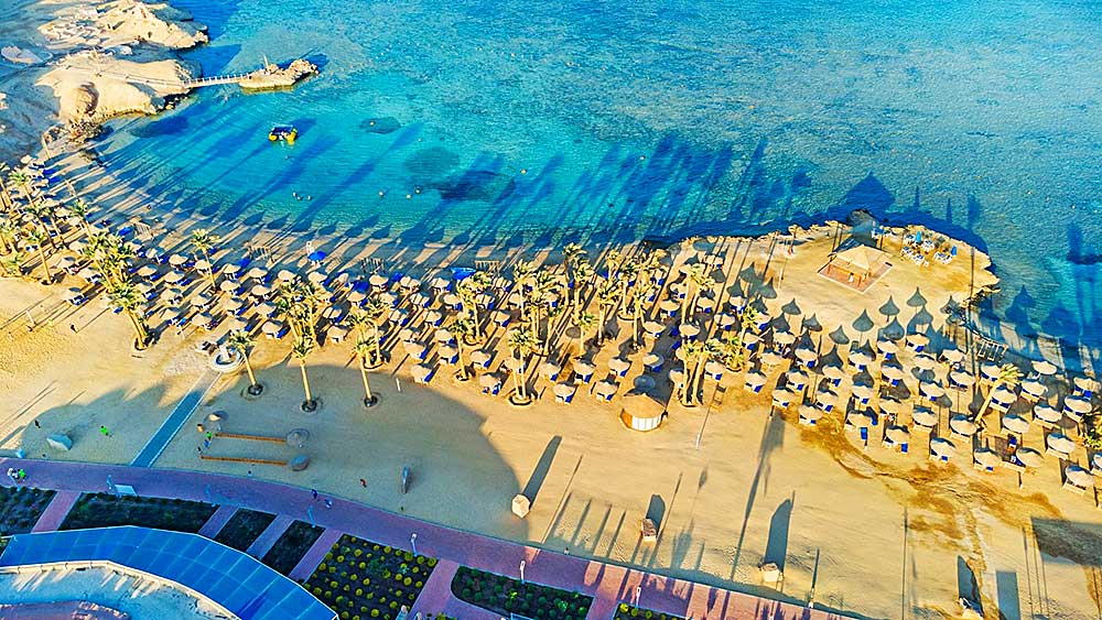 Una veduta aerea di una spiaggia tropicale soleggiata con file di ombrelloni e sedie a sdraio lungo la battigia, evidenziando l'offerta viaggio del Villaggio Mar Rosso.