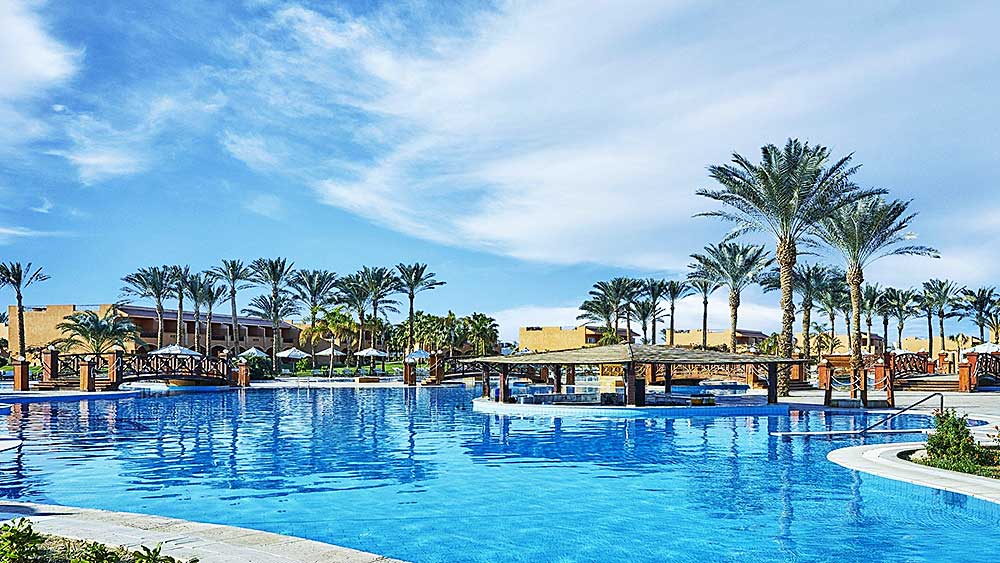 Una tranquilla piscina in stile Villaggio Mar Rosso con palme e lettini sotto un cielo azzurro e limpido.