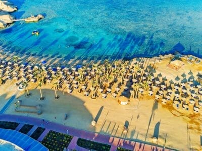 Veduta aerea di una soleggiata località balneare a Marsa Alam con ombrelloni che proiettano lunghe ombre sulla sabbia, accanto alle calme acque turchesi.