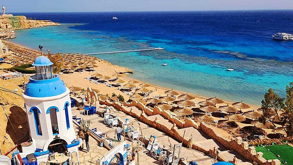 Una zona turistica costiera con una struttura a cupola blu che si affaccia su una spiaggia sabbiosa con ombrelloni e un mare limpido e turchese nel villaggio di Sharm El Sheikh.