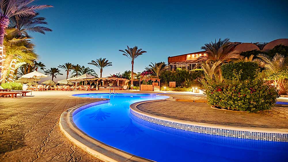 Piscina illuminata del Villaggio Mar Rosso al crepuscolo con palme e lettini circostanti.