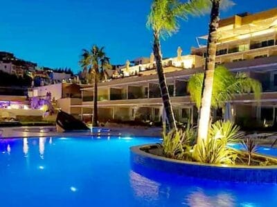 Vista notturna di una piscina illuminata con una piccola isola e palme di fronte a edifici bianchi ornati di luci in un hotel 4* a Muskebi.