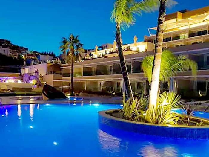 Vista notturna di una piscina illuminata con una piccola isola e palme di fronte a edifici bianchi ornati di luci in un hotel 4* a Muskebi.