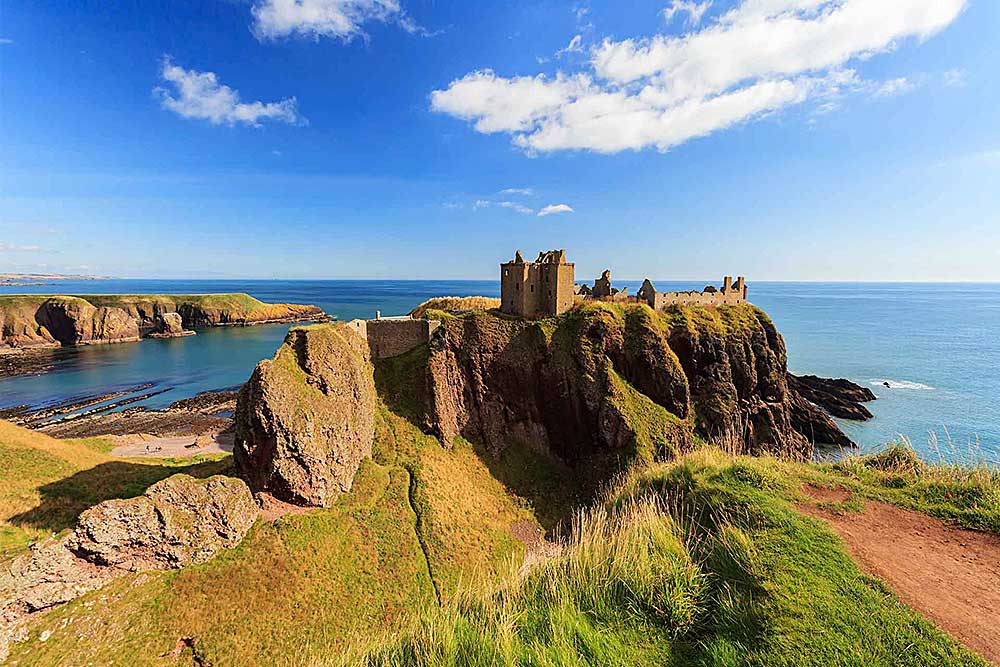 Le rovine di un castello si ergono su una scogliera erbosa a picco sull'oceano, con un cielo azzurro e nuvole sparse sullo sfondo. Scopri questo scenario incantevole con un'offerta viaggio in Scozia di SBS Viaggi, dove la storia incontra la bellezza naturale.