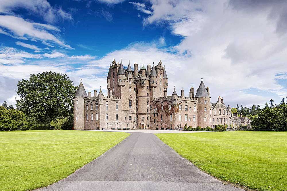 Un maestoso castello in pietra con molteplici torri e torrette si erge circondato da prati verdi sotto un cielo azzurro parzialmente nuvoloso, offrendo una vista pittoresca perfetta per un SBS Viaggi offerta viaggio in Scozia.