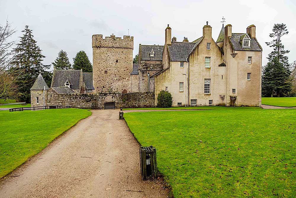 Uno storico castello in pietra con molteplici camini e torrette è circondato da prati verdi e alberi, con un sentiero di ghiaia che conduce all'ingresso. Scopri questa incantevole destinazione con l'offerta viaggio in Scozia di SBS Viaggi, trasformando la tua fuga da sogno in realtà.