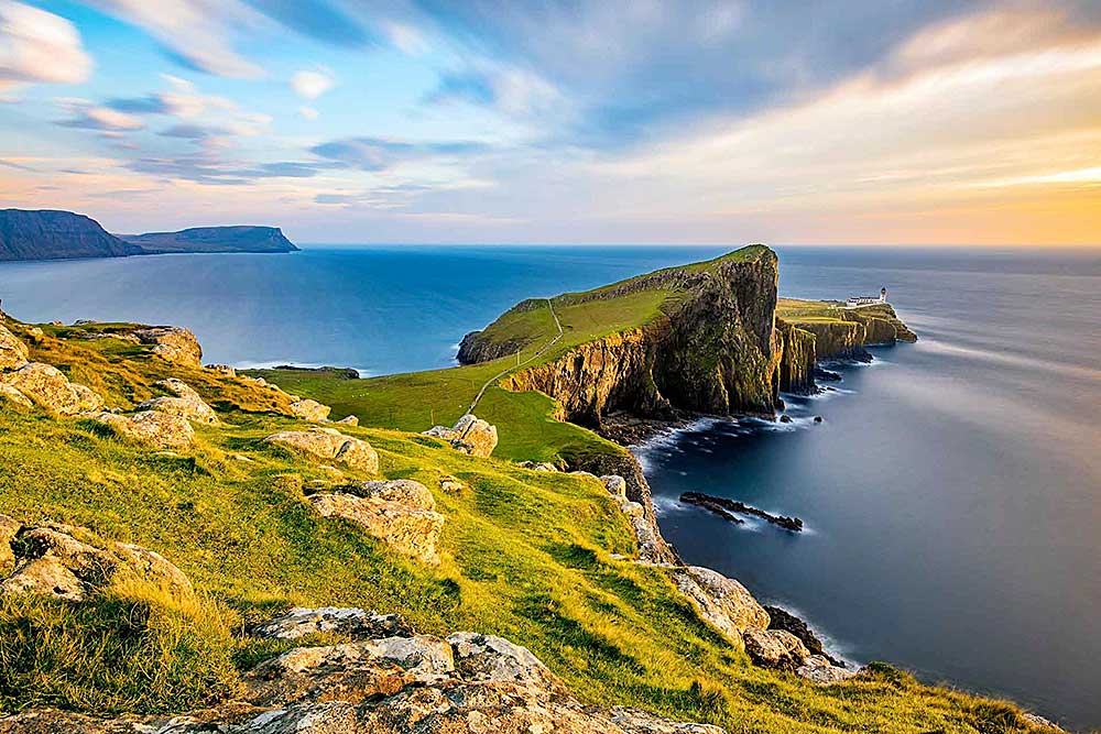 Un faro sulla scogliera si affaccia sull'oceano al tramonto, con colline erbose e una costa rocciosa. Il cielo ha nuvole sparse e una sfumatura di colori dall'azzurro all'arancione, catturando la magia di un'offerta viaggio in Scozia di SBS Viaggi.
