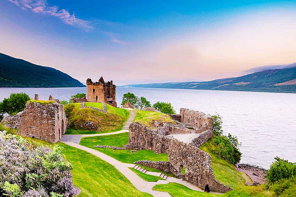 Un castello in rovina si trova su una collina erbosa accanto a un grande lago, con le montagne sullo sfondo sotto un cielo limpido. I sentieri si snodano attorno alle rovine del castello e al paesaggio: uno scenario perfetto per un viaggio in Scozia di SBS Viaggi.