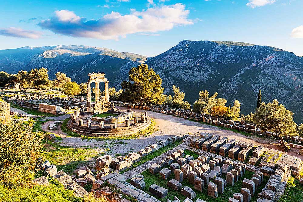 Scopri le antiche rovine del Santuario di Atena Pronaia a Delfi, in Grecia, con montagne e alberi circostanti sotto un cielo parzialmente nuvoloso. Prenota la tua prossima avventura con SBS Viaggi e approfitta della nostra esclusiva "Offerta viaggio in Grecia.