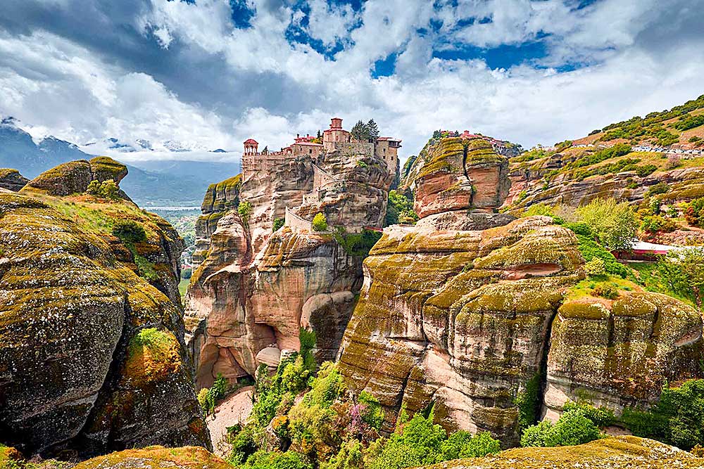 Uno storico monastero si trova in cima a imponenti formazioni rocciose sotto un cielo parzialmente nuvoloso, circondato da una vegetazione lussureggiante e aspre scogliere: vivi questa vista mozzafiato con un'Offerta viaggio in Grecia di SBS Viaggi.