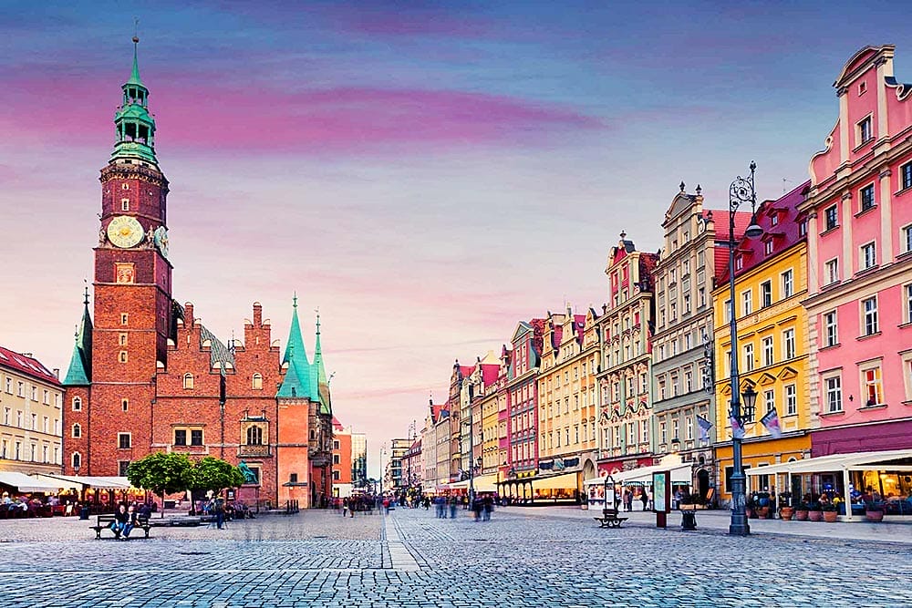 Una storica piazza cittadina con pavimentazione in ciottoli, edifici colorati e un'importante torre dell'orologio sotto un cielo rosa al tramonto. Le persone passeggiano e cenano nei caffè all'aperto. Vivi questo scenario affascinante con l'offerta viaggio in Polonia di SBS Viaggi.