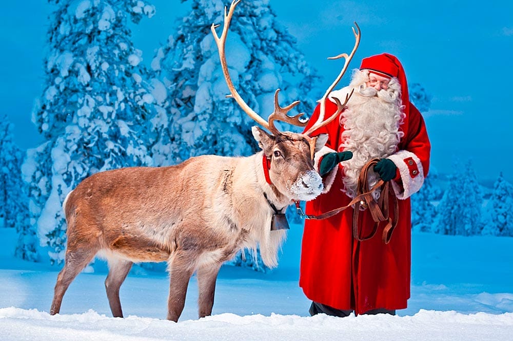 Una persona vestita da Babbo Natale sta in piedi nella neve, tiene le redini e accarezza una renna. Sullo sfondo ci sono alberi innevati: uno scorcio magico di ciò che ti aspetta con un'offerta viaggio in Lapponia di SBS Viaggi.