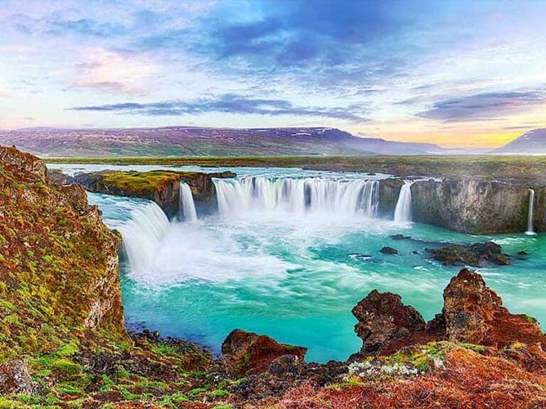 Una scenografica cascata precipita in un fiume turchese circondato da terreno roccioso e vegetazione muschiosa, offrendo un paesaggio pittoresco che potrebbe facilmente far parte di un Gran Tour dell'Islanda, con un cielo nuvoloso sullo sfondo.