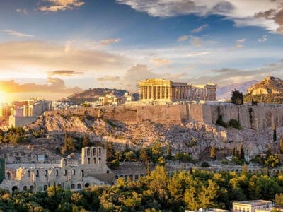 Veduta dell'Acropoli di Atene, Grecia, con il Partenone in cima a uno sperone roccioso circondato da antiche strutture, alberi e uno spettacolare cielo al tramonto.