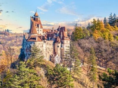 Un castello medievale con torri appuntite si trova in cima a una collina rocciosa, circondato da alberi autunnali sotto un cielo limpido, evocando una scena uscita da una favola rumena.